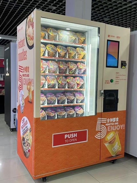 Vending machine stocked with packaged food items, featuring a glass display front, touchscreen interface, and orange base with push-to-open instructions.