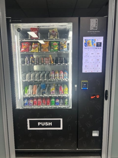 Black vending machine with illuminated glass front displaying beverages and snacks, digital menu screen, and PUSH button
