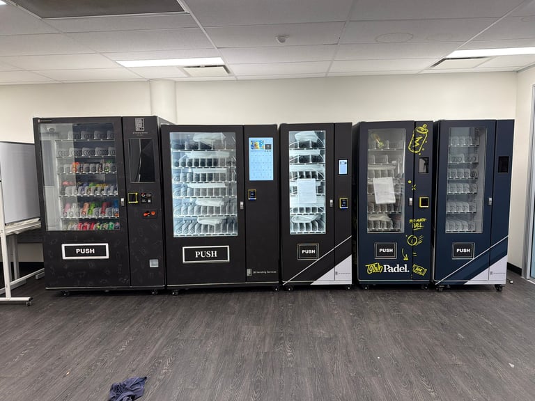 Row of black vending machines in an office or break room displaying snacks and beverages with illuminated product shelves